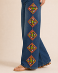 Close-up of a person wearing blue wide-leg jeans with four large colorful embroidered geometric diamond patterns down the outer legs, paired with tan sandals against a plain beige studio background.