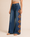 A model wearing high-waisted, wide-leg blue denim jeans featuring colorful embroidered geometric diamond patterns down the outer side of the legs, paired with tan sandals against a solid beige studio background.