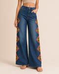 A model wearing blue wide-leg denim jeans with colorful embroidered geometric patterns down the outer sides, paired with a beige crop top and flat sandals, set against a solid light beige background.