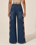 A model shown from the back wearing dark blue wide-leg denim jeans with colorful embroidered geometric patterns down the outer sides, set against a plain beige studio background.