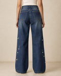 Back view of a model wearing high-waisted, wide-leg blue denim jeans with small embroidered floral details down the sides, paired with a white top against a warm beige studio background.