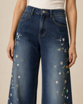 A person wearing dark blue wide-leg denim jeans with colorful floral embroidery down the front, paired with a white ribbed top against a plain beige studio background.