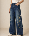 A person wearing high-waisted wide-leg blue denim jeans with colorful floral embroidery down the outer legs, paired with a white top and white sneakers, against a plain beige studio background.