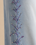 A model wearing light-wash wide-leg jeans with purple floral embroidery down the side, paired with a white top and white sneakers, against a solid beige background.