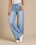 A model wearing light-wash wide-leg jeans with purple floral embroidery down the side, paired with a white top and white sneakers, against a solid beige background.