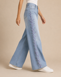 A model wearing light-wash wide-leg jeans with purple floral embroidery down the side, paired with a white top and white sneakers, against a solid beige background.