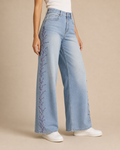 A model wearing light-wash wide-leg jeans with purple floral embroidery down the side, paired with a white top and white sneakers, against a solid beige background.